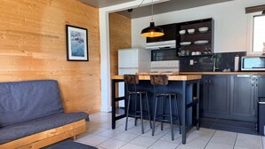 A modern kitchen with a wooden wall, a dining area with stools, a sofa, and a refrigerator. - Attitude Montagne (St-Adolphe-d'Howard)