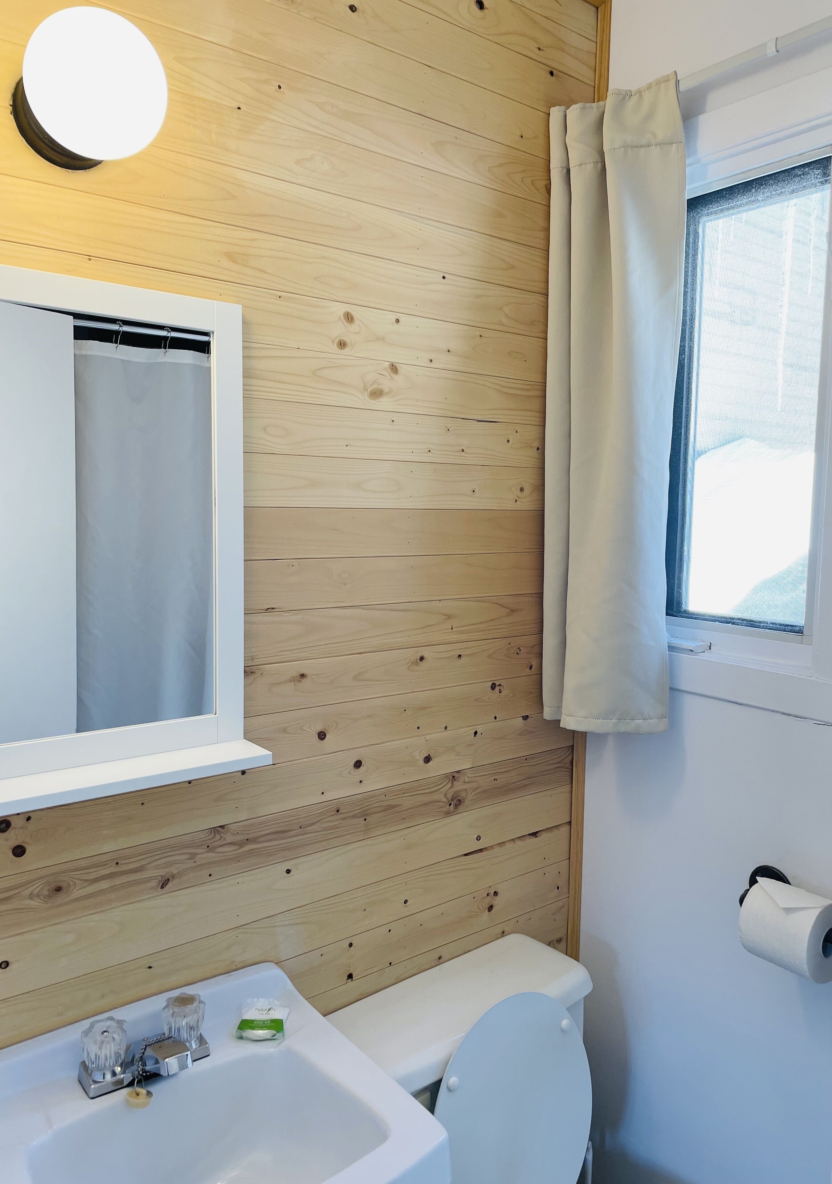 A bathroom with wooden panel walls, a white sink with glass soap dispensers, a toilet, and a window with a curtain.