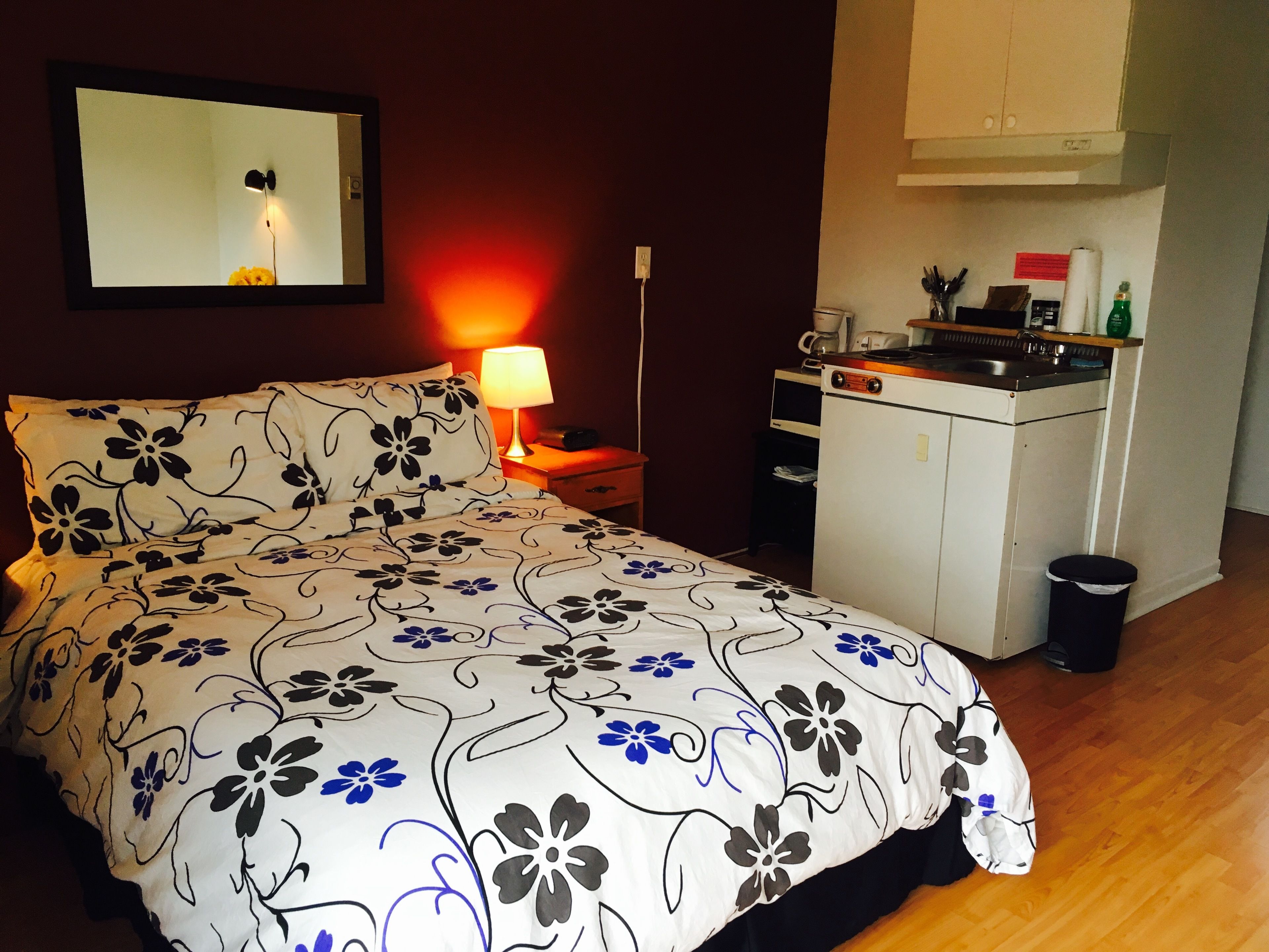A bedroom with a bed, a nightstand, a lamp, a mirror, a kitchenette, and a trash bin.