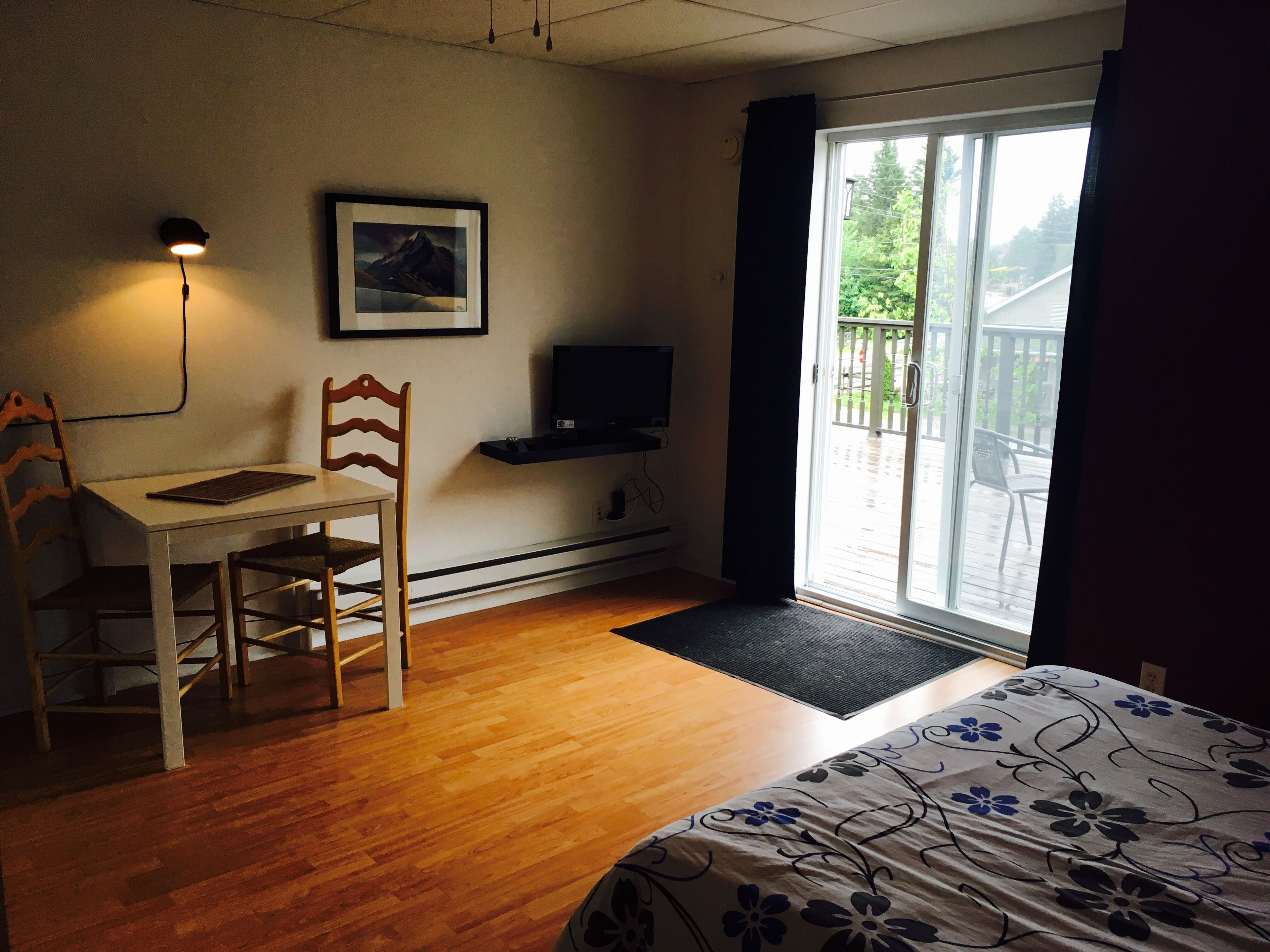 Une pièce comprenant un bureau en bois, une chaise, un lit recouvert d’une couvre-lit à motifs floraux, un téléviseur fixé au mur, un tableau encadré accroché au mur et une porte-fenêtre coulissante donnant sur un espace extérieur.