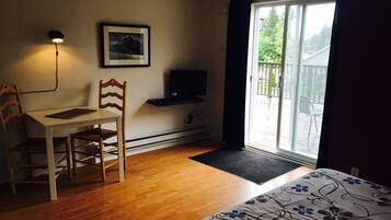 Une pièce comprenant un bureau en bois, une chaise, un lit recouvert d’une couvre-lit à motifs floraux, un téléviseur fixé au mur, un tableau encadré accroché au mur et une porte-fenêtre coulissante donnant sur un espace extérieur.