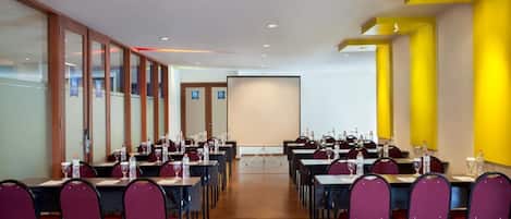 Un salón de conferencias con varias mesas y sillas, equipado con botellas de agua y vasos, y una pantalla de proyector al frente.