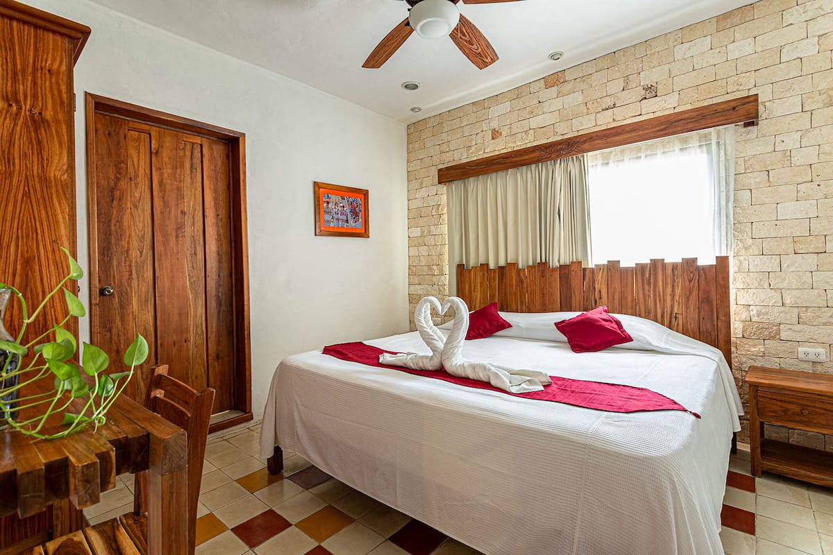 Habitación de hotel con una cama grande, cabecera de madera, ventilador de techo, pared de ladrillo y ventana con cortinas.