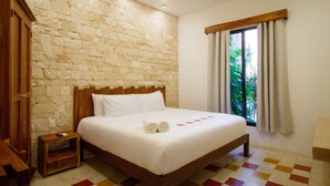 A neatly made bed with white linens and rolled towels, a wooden headboard, a stone wall, a window with curtains, and a checkered floor. - Hotel Casa Hipil (Valladolid)