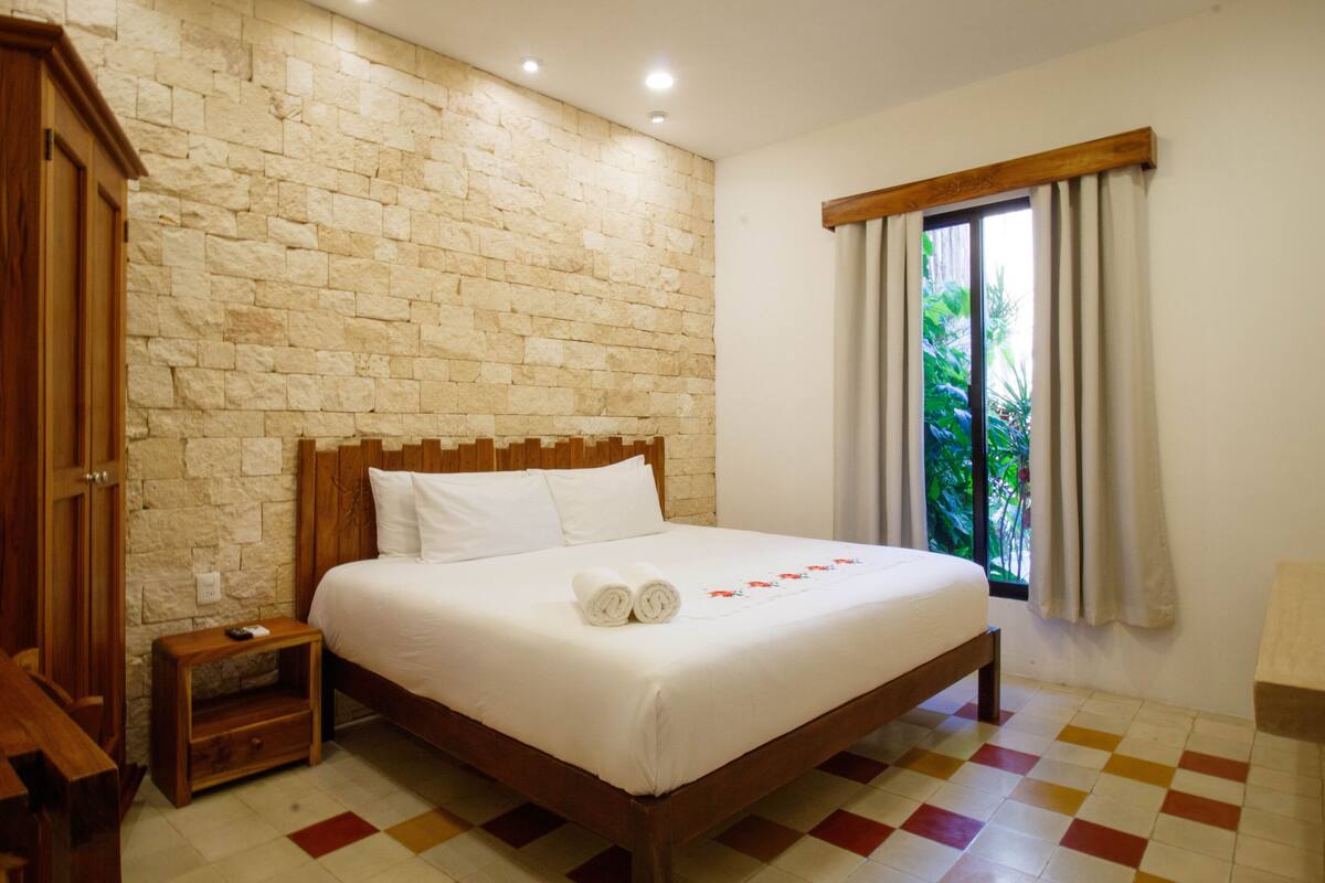 Una cama bien hecha con sábanas blancas y toallas enrolladas, cabecera de madera, pared de piedra, ventana con cortinas y suelo a cuadros.