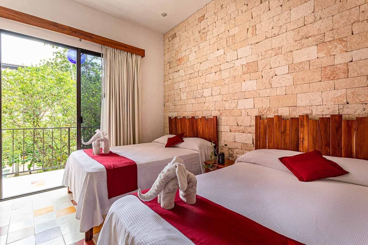 Habitación de hotel con dos camas, una pared de ladrillo y un balcón con vistas a zonas verdes.
