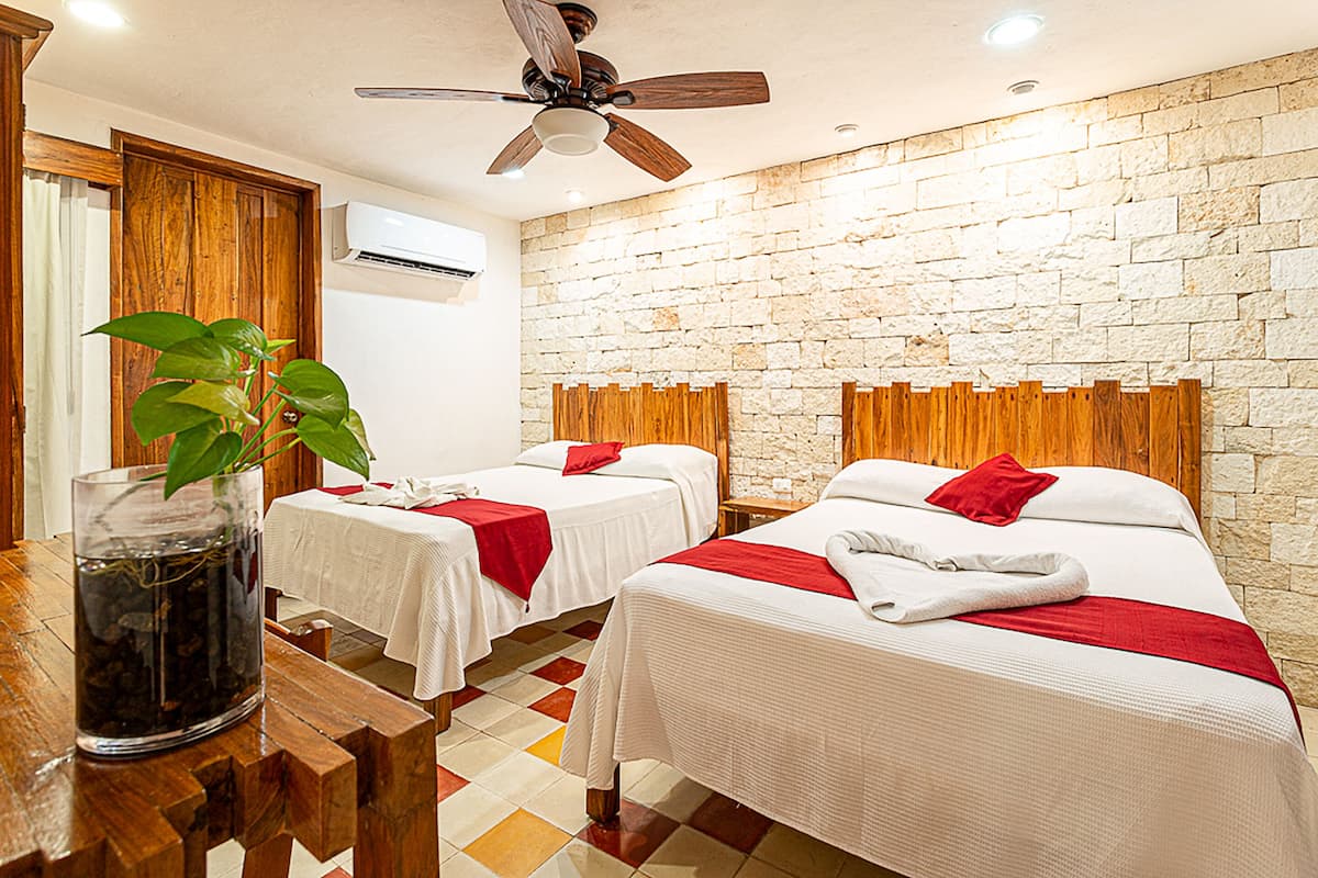 Habitación de hotel con dos camas, ventilador de techo de madera, pared de piedra y piso de baldosas.