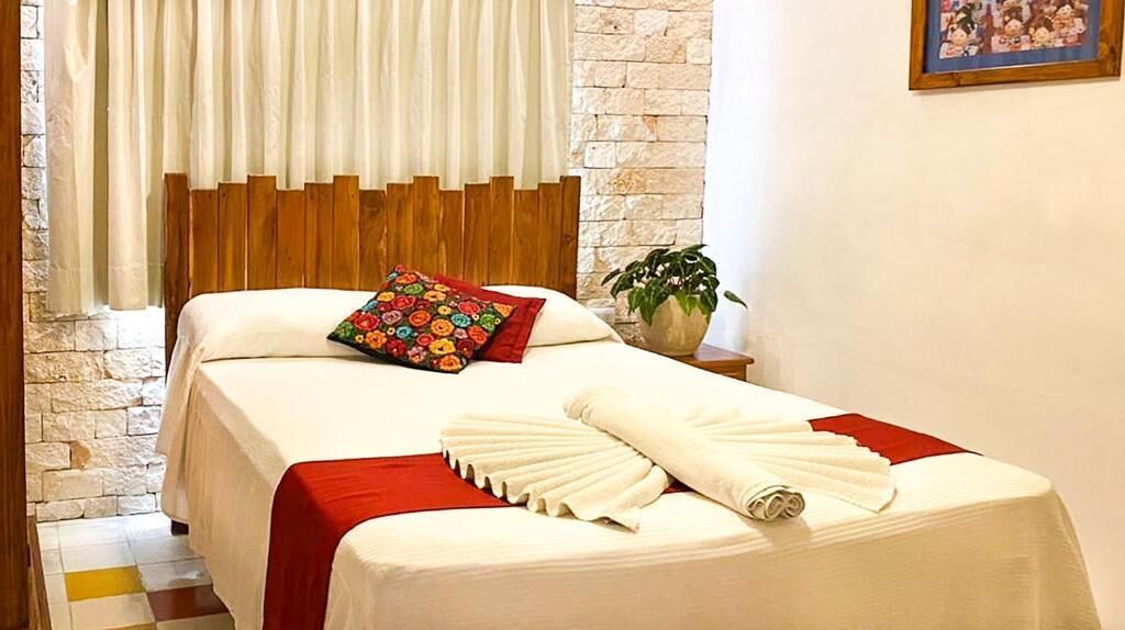 Camera d'albergo con un letto, una testiera in legno, una coperta a letto rossa e bianca, un cuscino colorato, un asciugamano bianco, un tavolino con una pianta e un quadro incorniciato appeso al muro.
