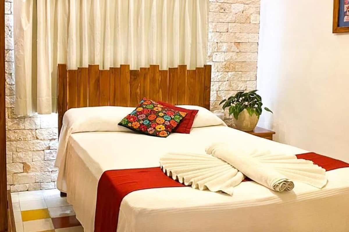 Habitación de hotel con cama, cabecera de madera, colcha roja y blanca, almohada colorida, toalla blanca, una mesita con una planta y un cuadro en la pared.