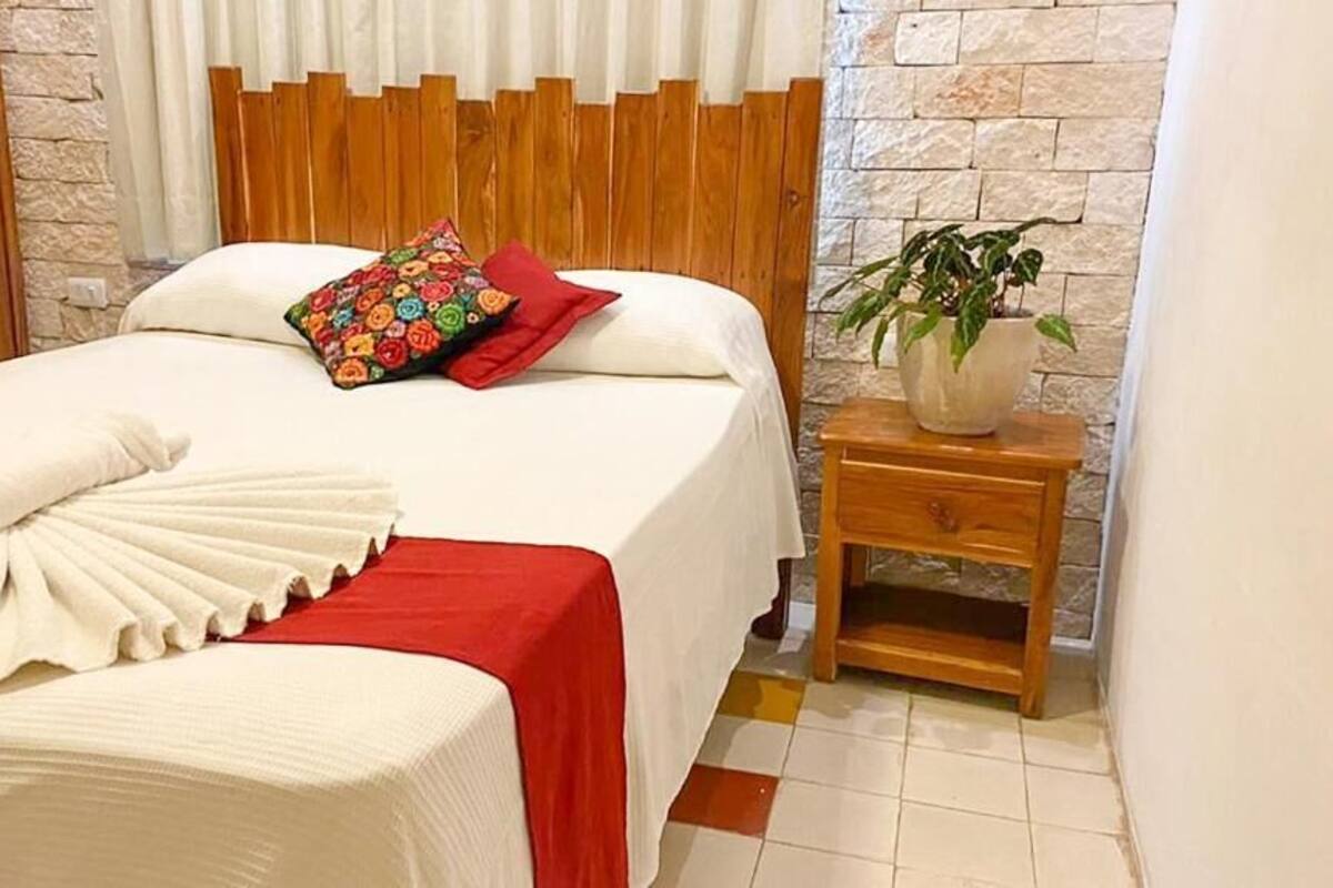 Una cama bien hecha con colcha roja y blanca, una almohada colorida y un cabecero de madera. Junto a la cama hay una mesita de noche de madera con una planta en maceta.