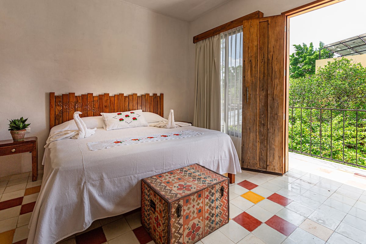 Un dormitorio con cabecera de madera, cama con ropa de cama blanca, una mesita con una planta y un otoman colorido.