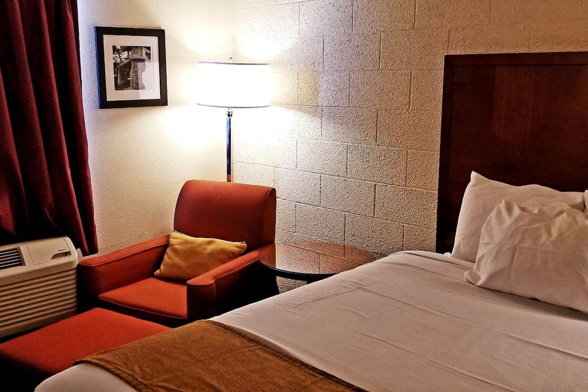 A hotel room with a bed, a red armchair, a lamp, and a picture on the wall.