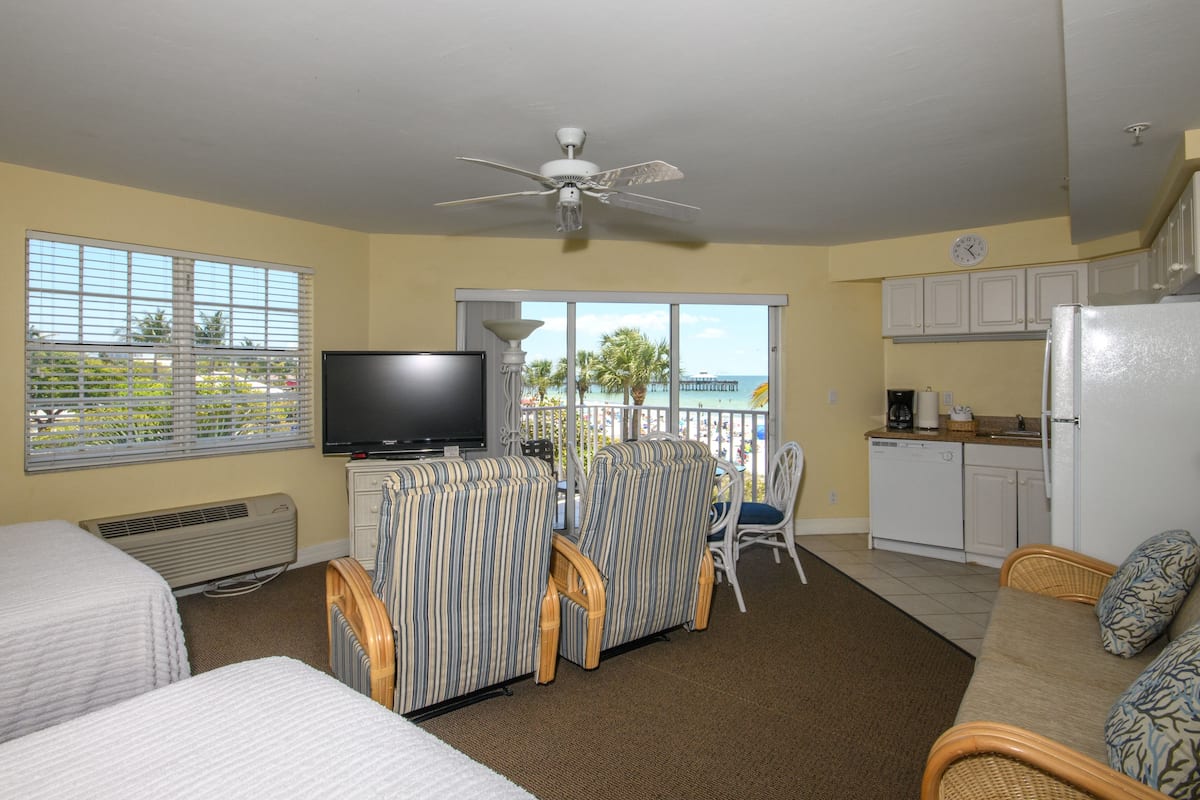 A hotel room with a TV, two beds, a dining area with chairs, a kitchen, and a balcony with a view of the beach.