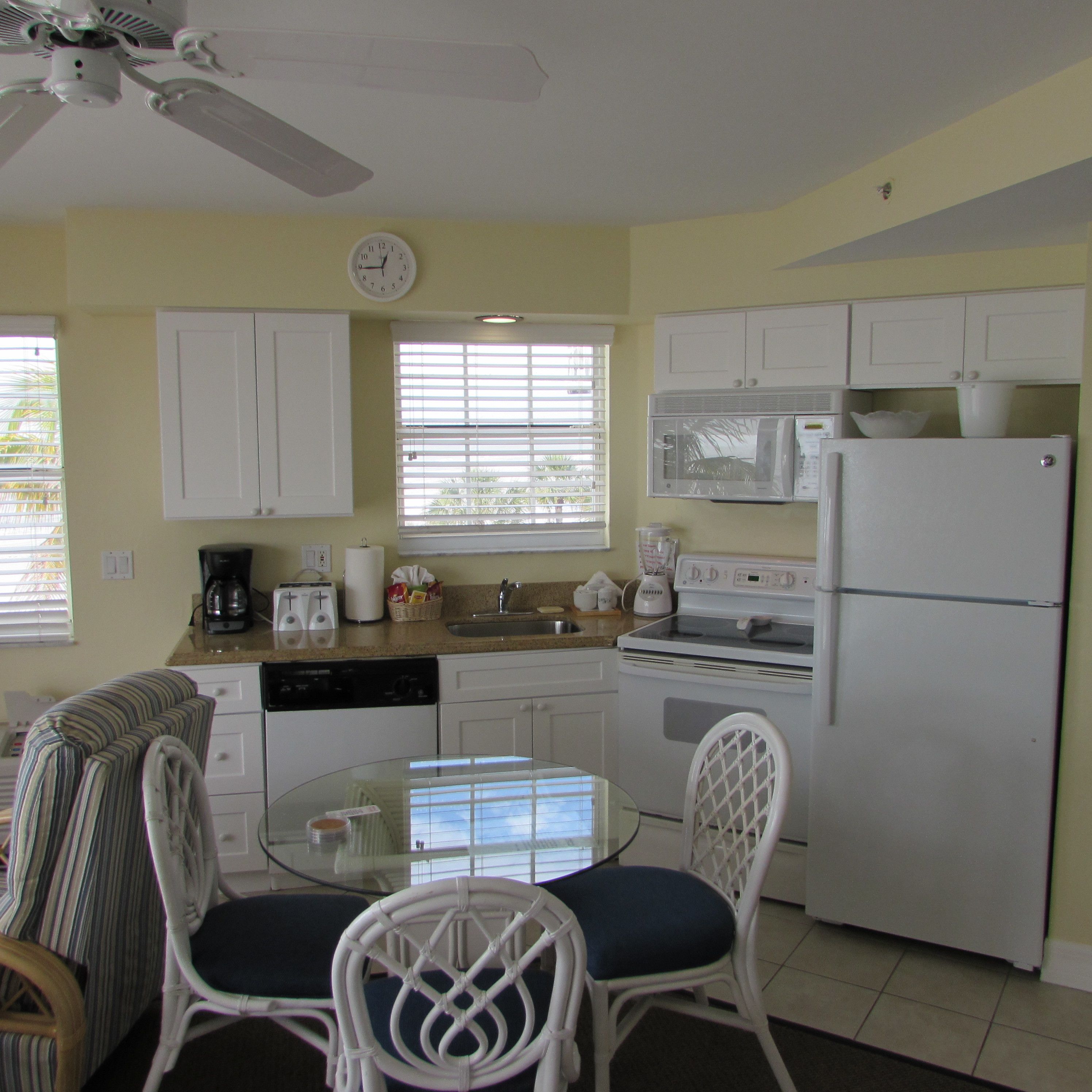 A compact kitchen with white appliances, a dining area with a glass table and wicker chairs, and a window with blinds.