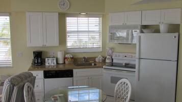 A compact kitchen with white appliances, a dining area with a glass table and wicker chairs, and a window with blinds.