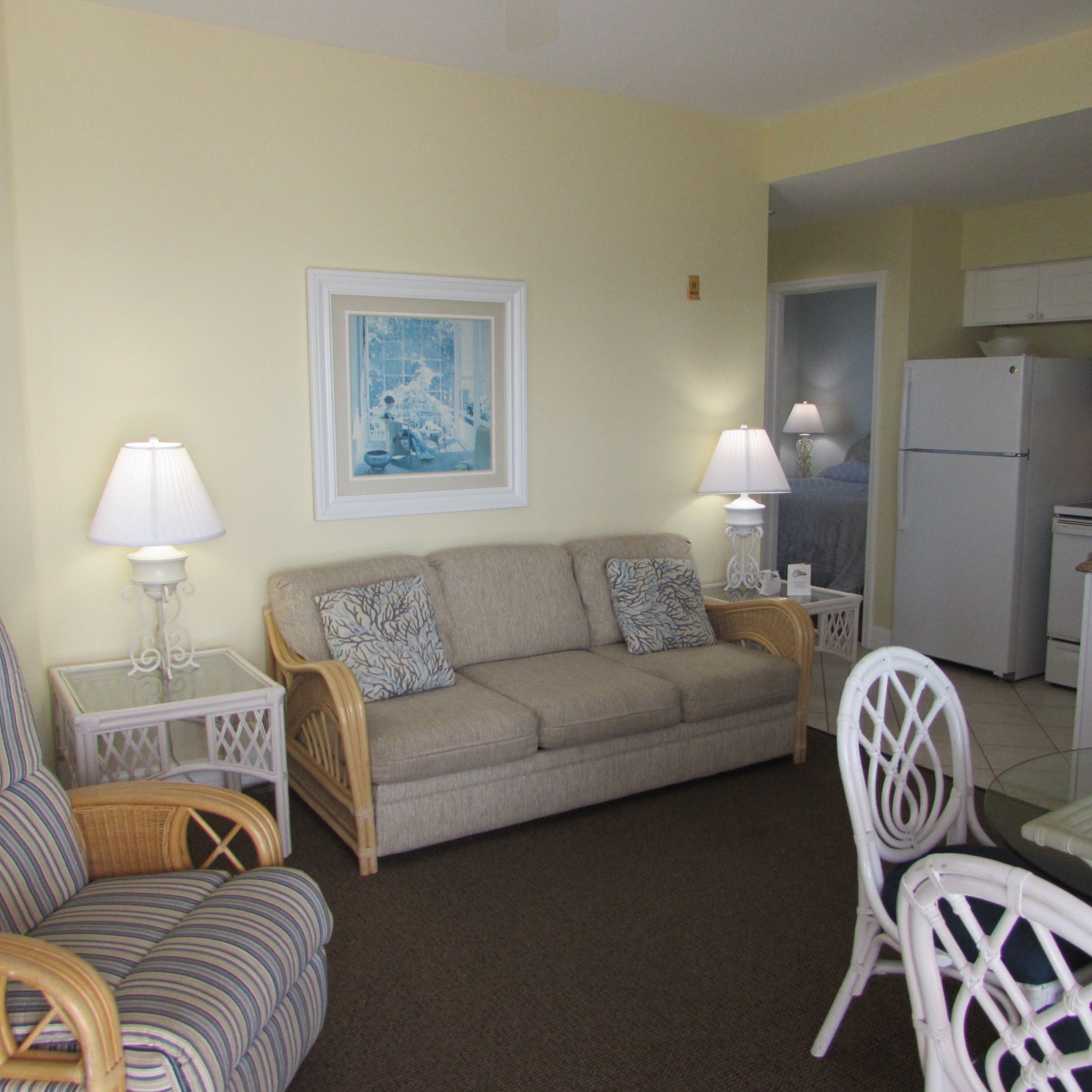 A living room with a sofa, two side tables with lamps, a framed picture on the wall, and a kitchen area with a refrigerator.