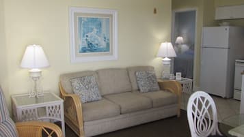 A living room with a sofa, two side tables with lamps, a framed picture on the wall, and a kitchen area with a refrigerator.