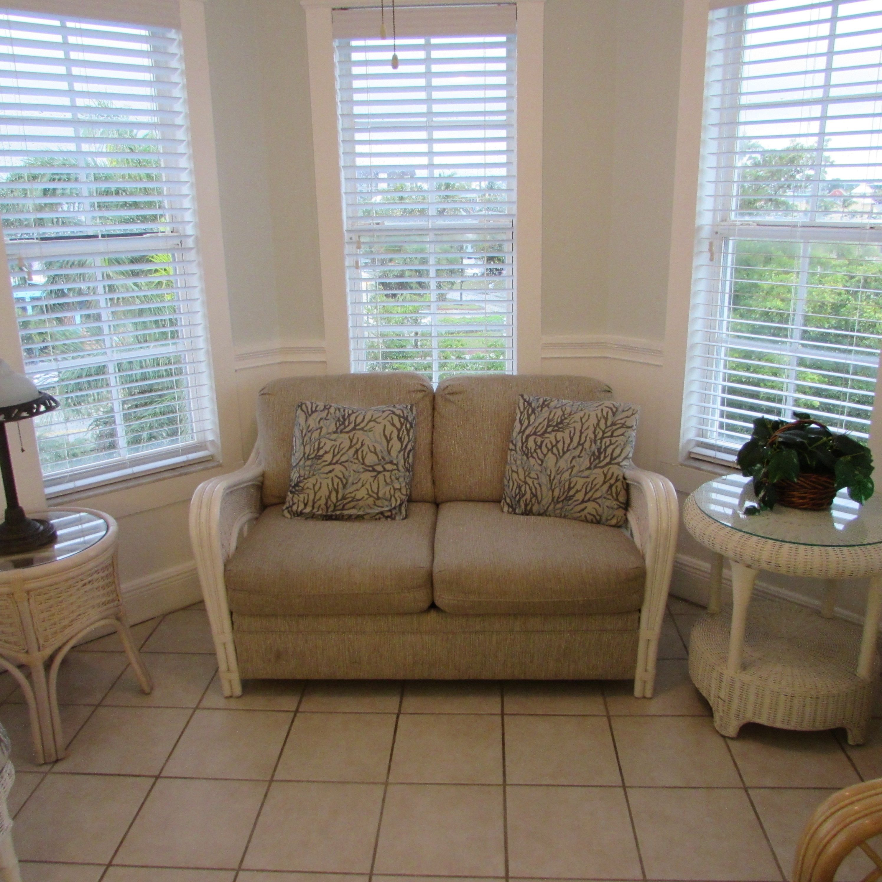 A beige sofa with two patterned pillows, a wicker side table, and a glass table with a plant.