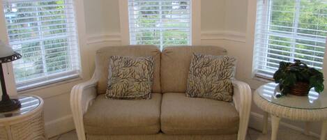 A beige sofa with two patterned pillows, a wicker side table, and a glass table with a plant.