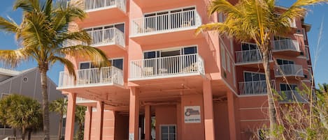 A multi-story hotel with balconies, a sandy ground floor, and palm trees.