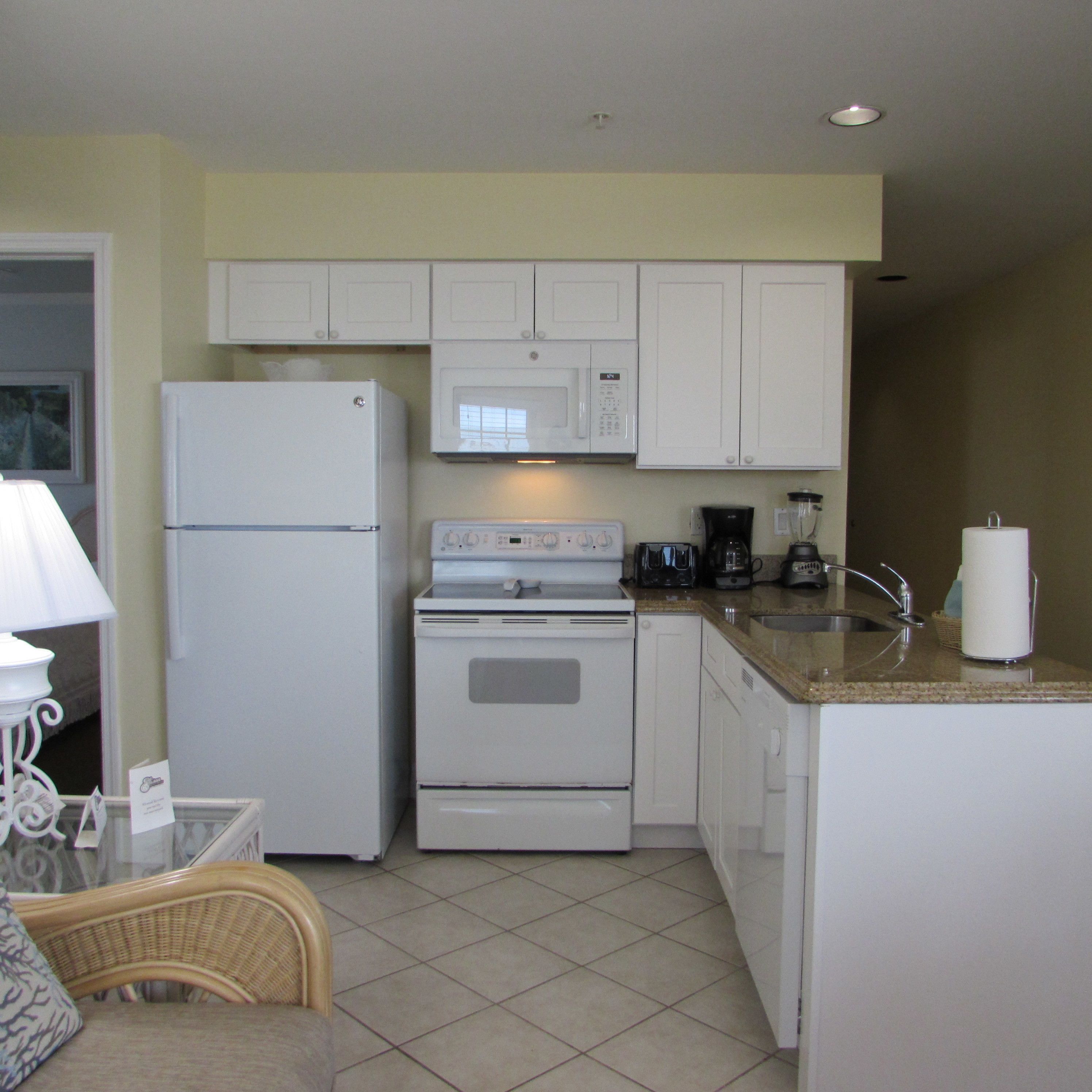 A compact kitchen with white appliances, including a refrigerator, microwave, and stove. There is a sink with a faucet, a coffee maker, and a blender on the countertop. The kitchen has white cabinets and a tiled floor.