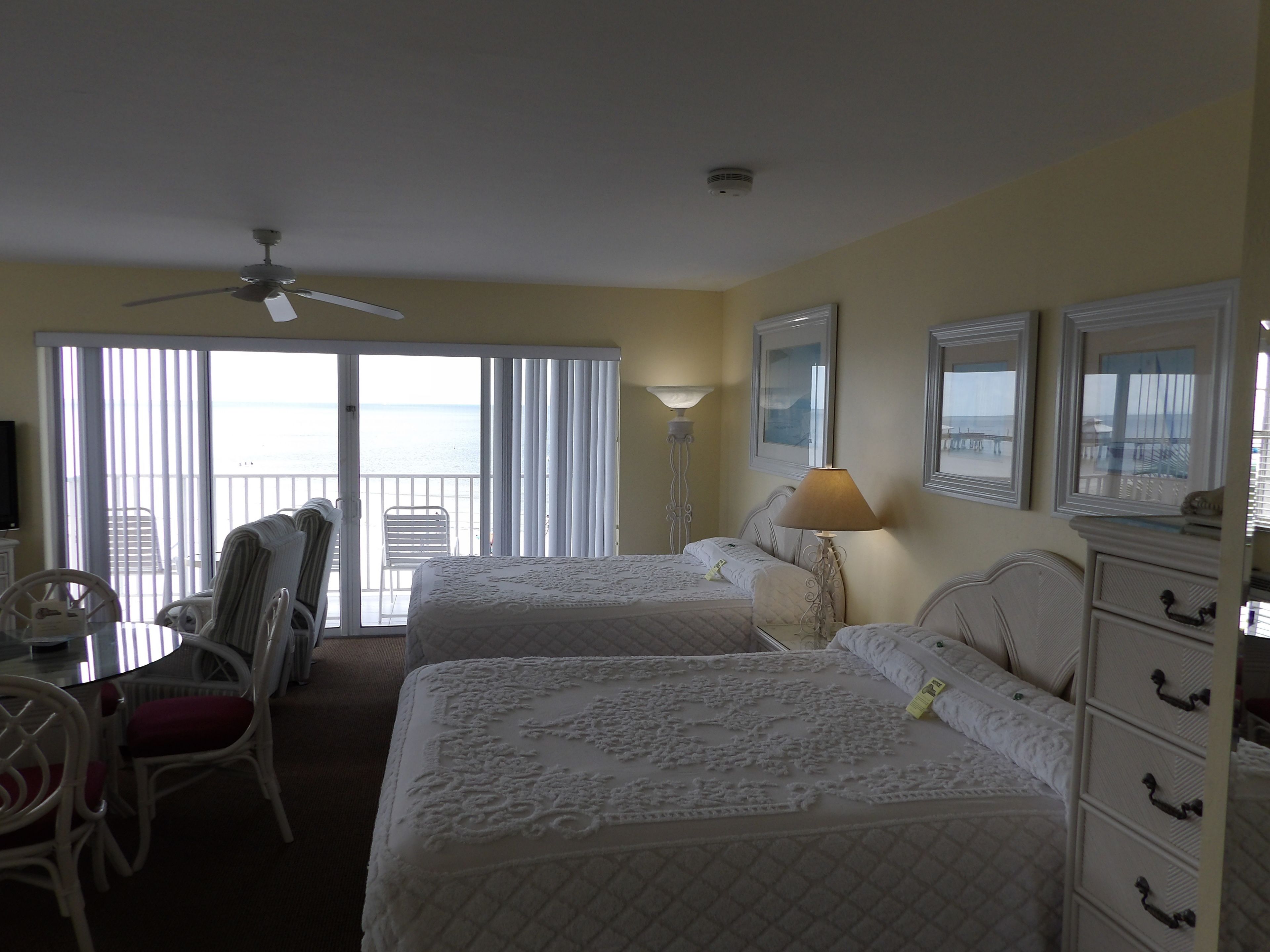 Une chambre d’hôtel avec deux lits, un ventilateur de plafond, un balcon avec vue sur la mer et un coin repas avec une table et des chaises.