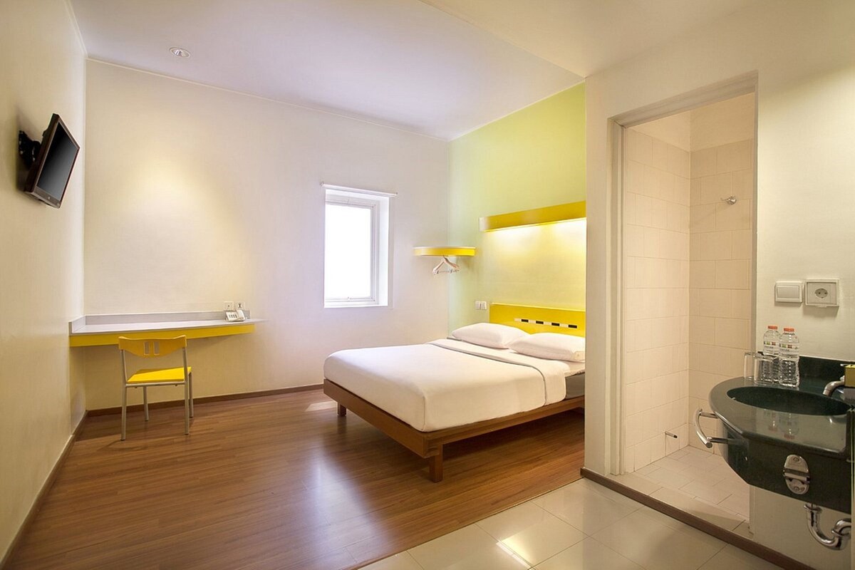 Quarto de hotel com cama, escrivaninha com cadeira, televisão na parede, janela e banheiro com pia e espelho.