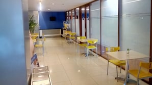 Um espaço amplo e interno, com mesas e cadeiras amarelas e brancas, uma parede azul e uma janela grande.