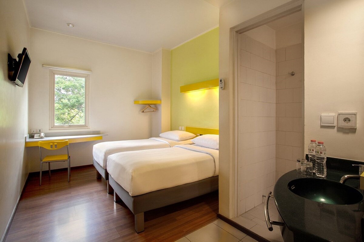 Quarto de hotel com duas camas, uma escrivaninha, uma cadeira, uma janela e banheiro com pia e espelho.