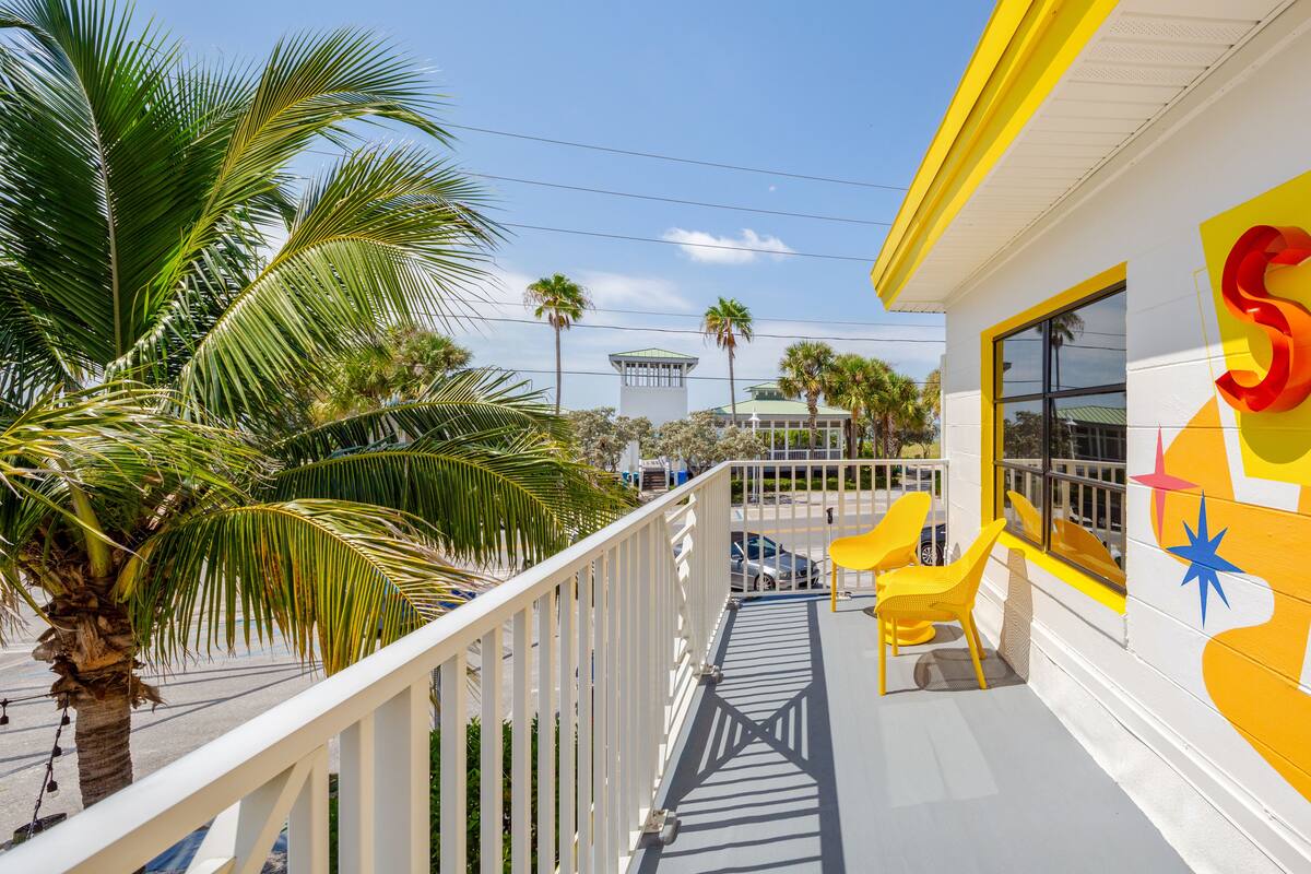A balcony with a white railing, yellow chairs, and a view of palm trees and a building with a green roof.