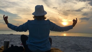 Eine Person sitzt auf einer Decke am Strand und blickt auf den Sonnenuntergang.