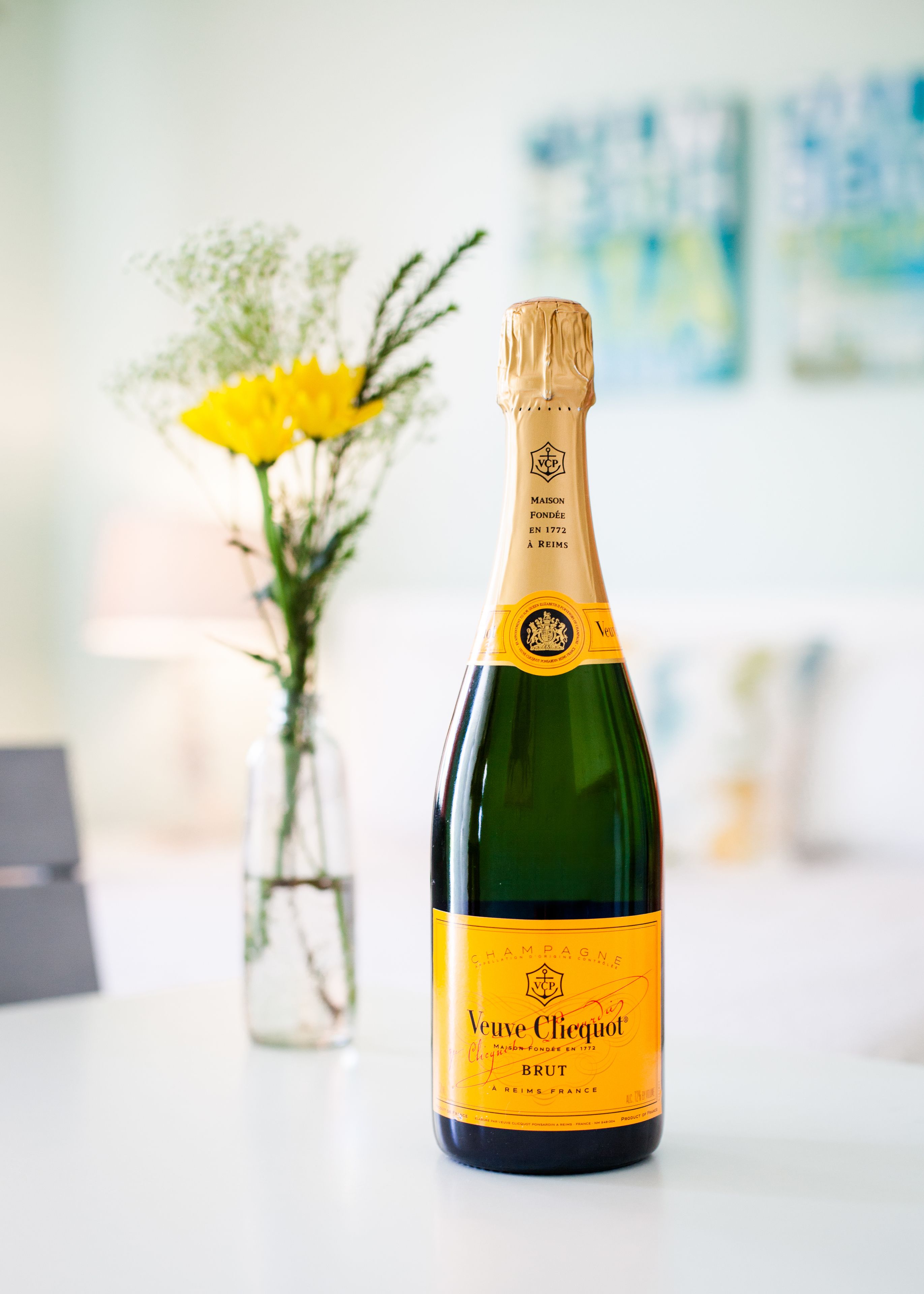Una botella de champán Veuve Clicquot Brut con etiqueta amarilla, sobre una mesa con un jarrón de flores amarillas al fondo.