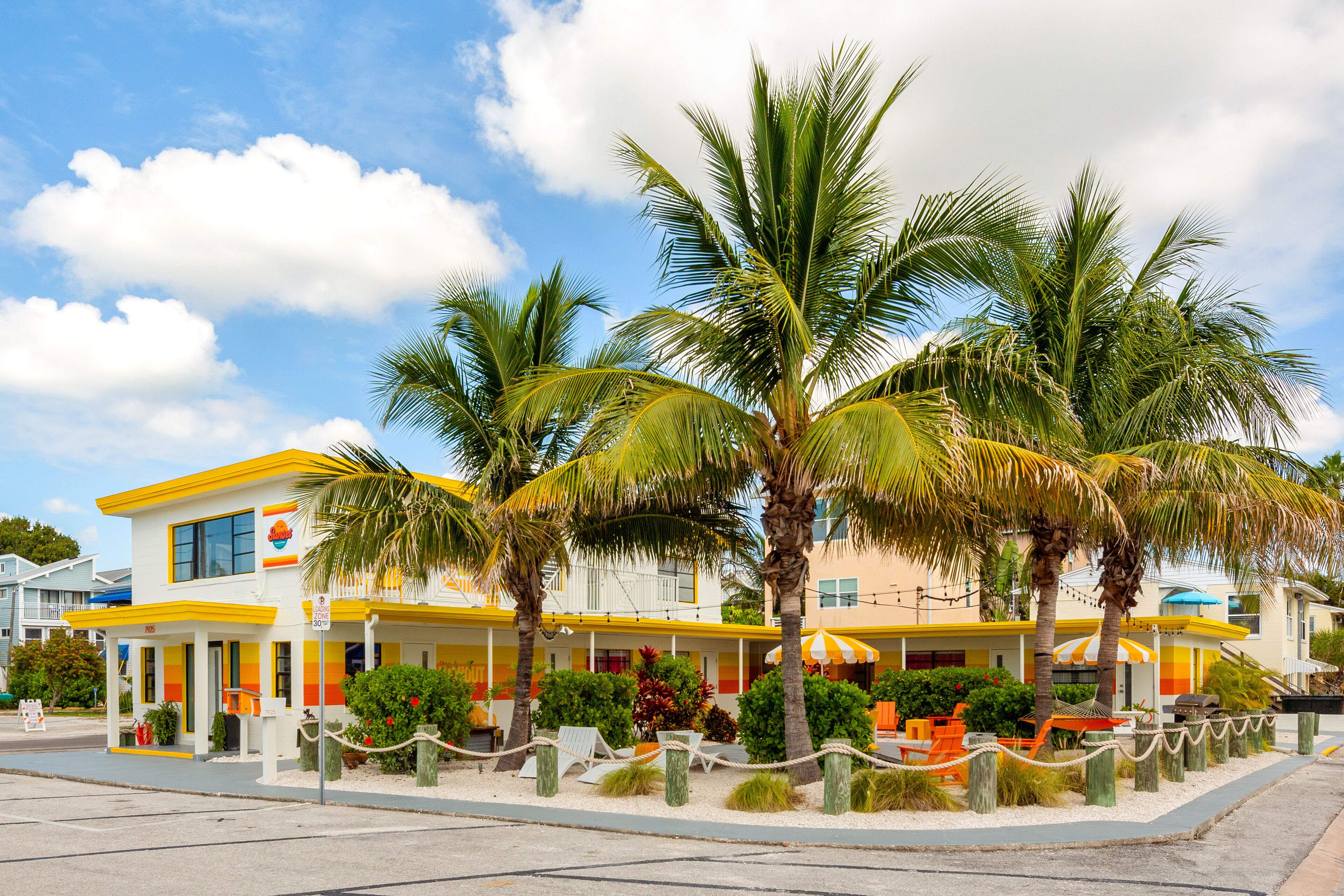 Un hotel con fachada amarilla y blanca, palmeras y mesas al aire libre.