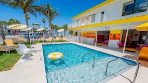 Ein Schwimmbecken mit Liegestühlen, einem Rettungsschwimmerstand und einem Gebäude mit gelb-weißer Fassade.