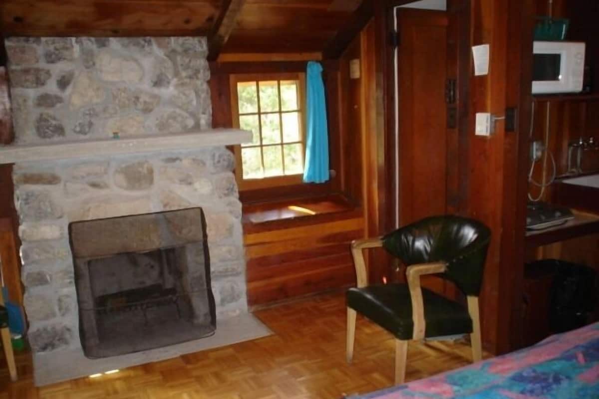 A rustic cabin interior with a stone fireplace, wooden walls, a window with a blue curtain, and a wooden chair.
