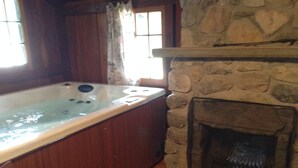 A stone fireplace with a built-in wood-burning stove, a wooden bathtub, and a window with curtains. - Addison's Bungalows (Brisco)