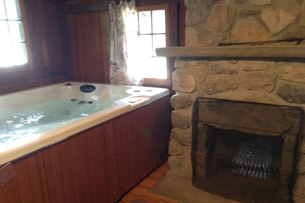 A stone fireplace with a built-in wood-burning stove, a wooden bathtub, and a window with curtains.