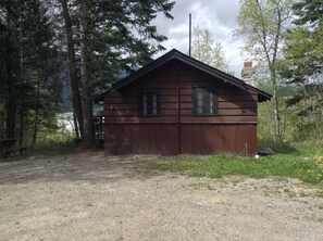A wooden cabin with two windows, surrounded by trees and a gravel path.