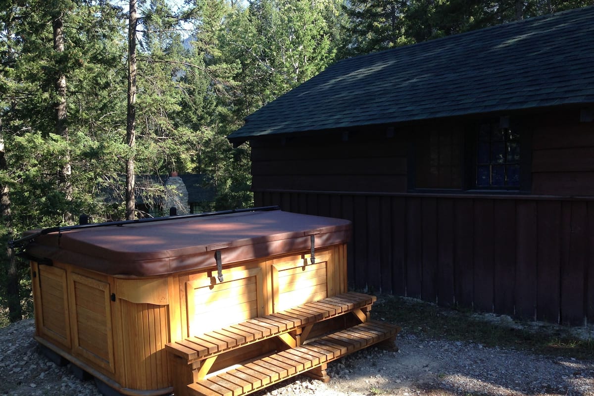 A wooden hot tub with steps, surrounded by a gravel area and a wooden building with a dark roof.