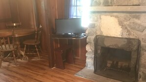 A cozy living room with a stone fireplace, wooden flooring, a dining table with chairs, and a television. - Addison's Bungalows (Brisco)