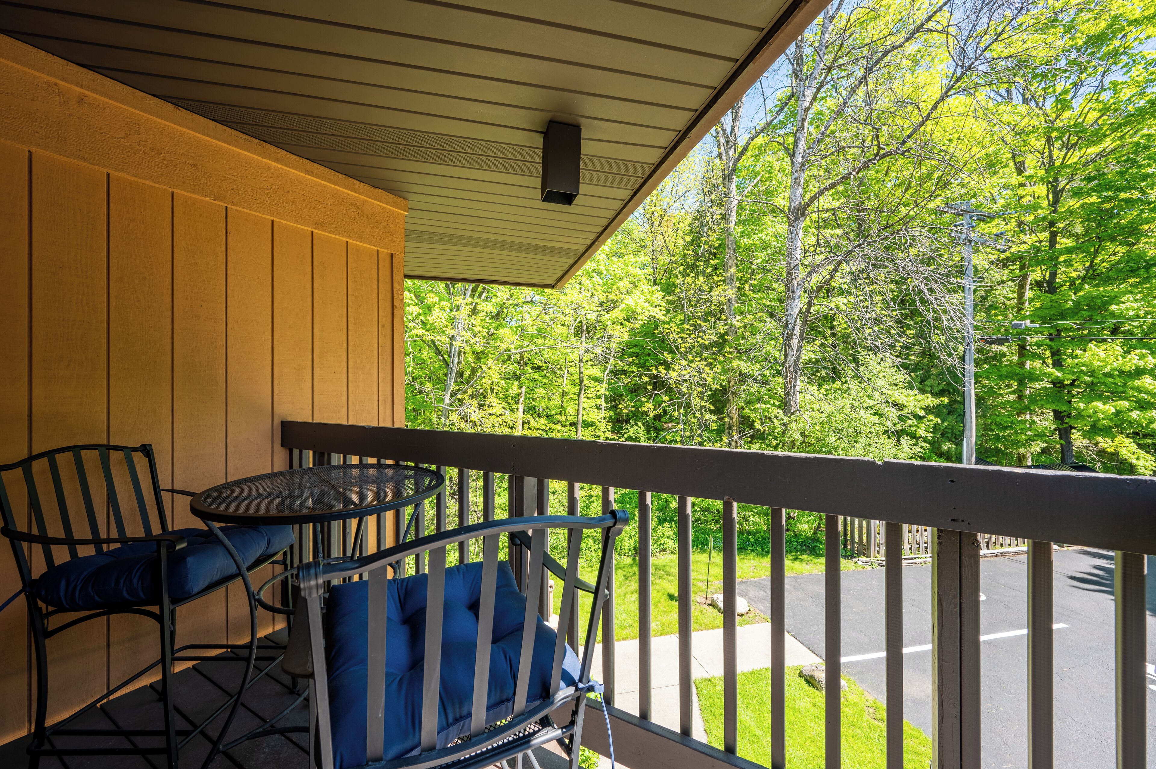 A covered outdoor seating area with a table and chairs, overlooking a road and greenery.
