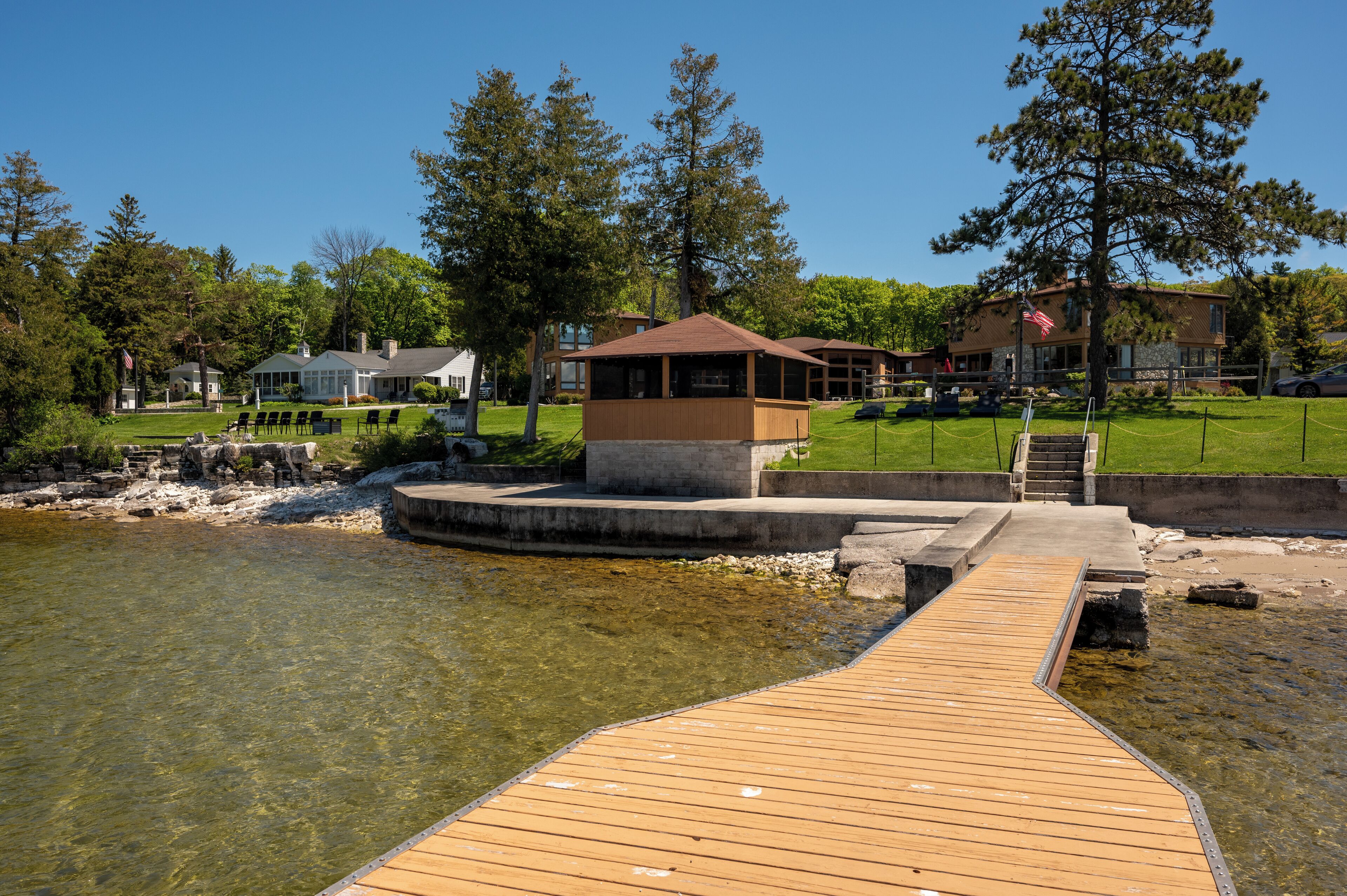 A wooden dock leading to a gazebo by a clear lake, surrounded by trees and a building with a flag.