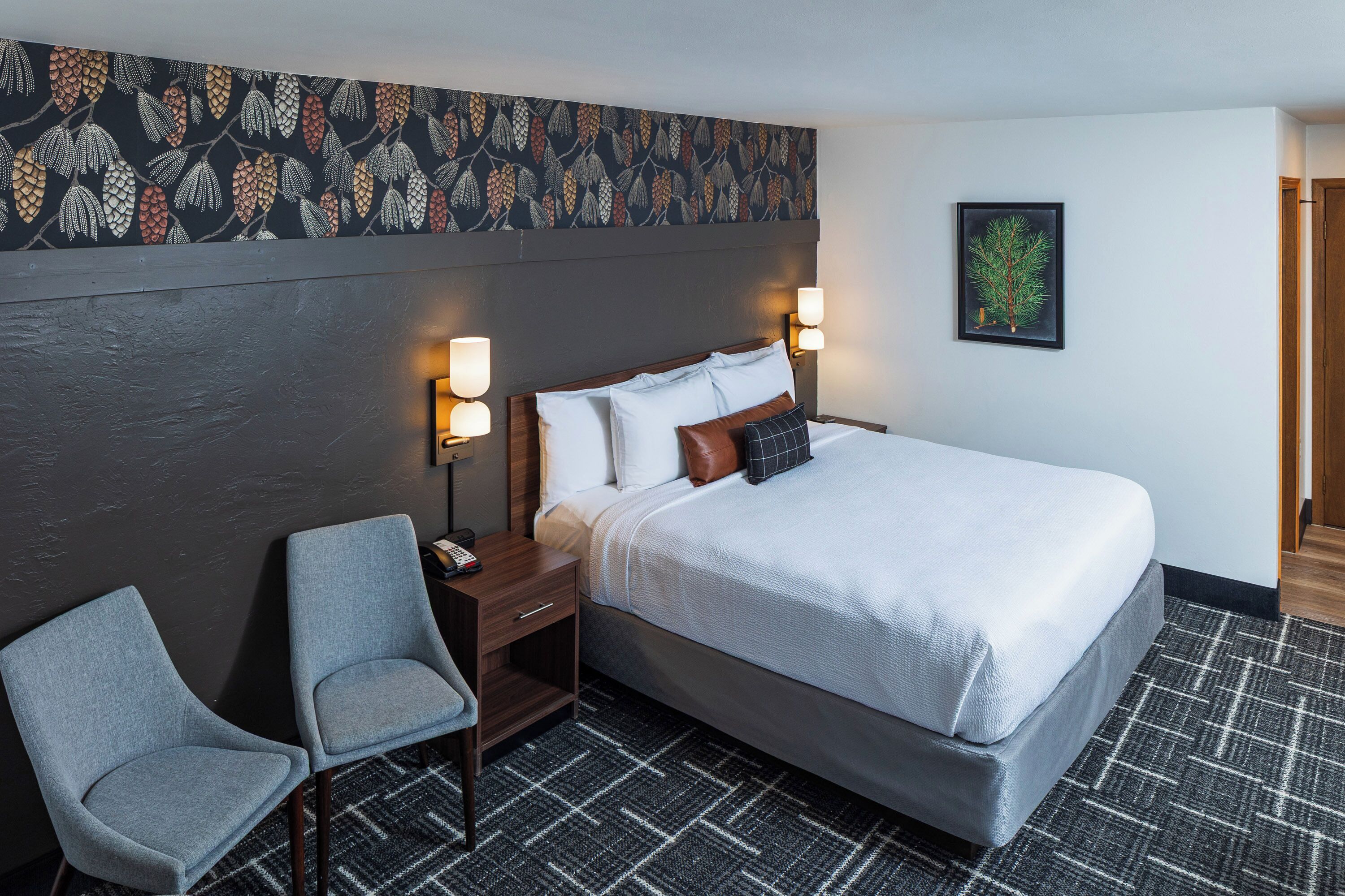 Une chambre d’hôtel comprenant un lit, deux lampes de chevet, une chaise, une table de chevet, une moquette à motifs et un mur décoré d’un motif de feuilles.