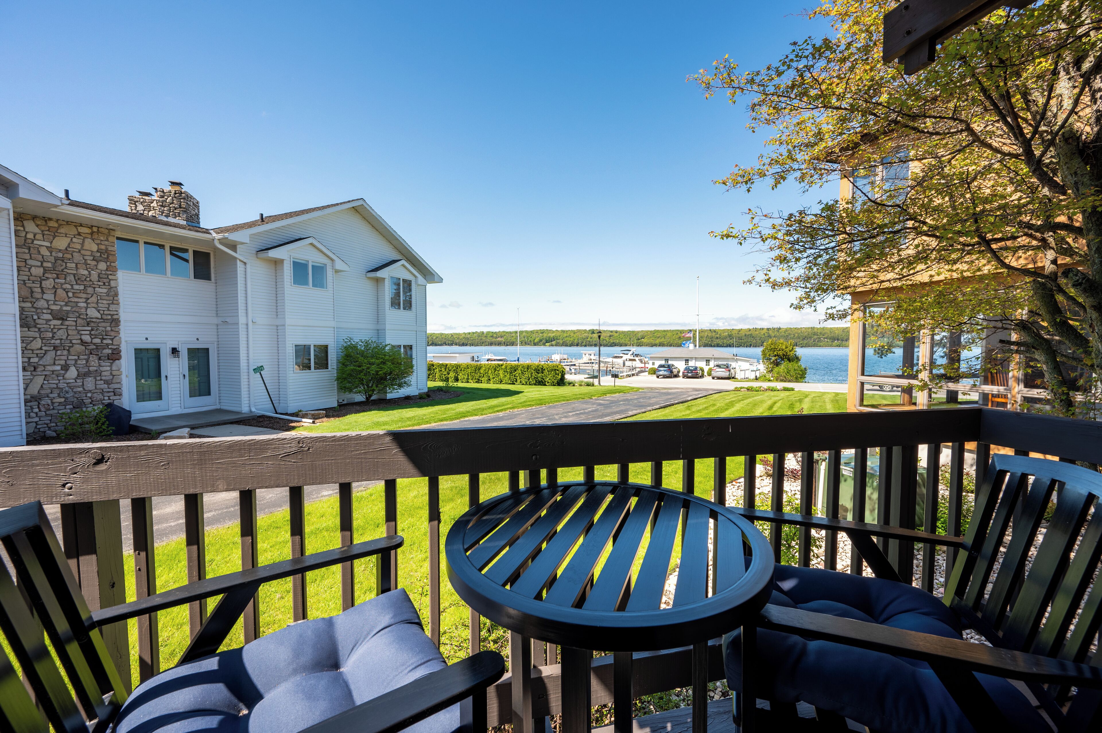 Une terrasse avec une table et des chaises, donnant sur un lac et une maison aux murs en pierre et au revêtement blanc.