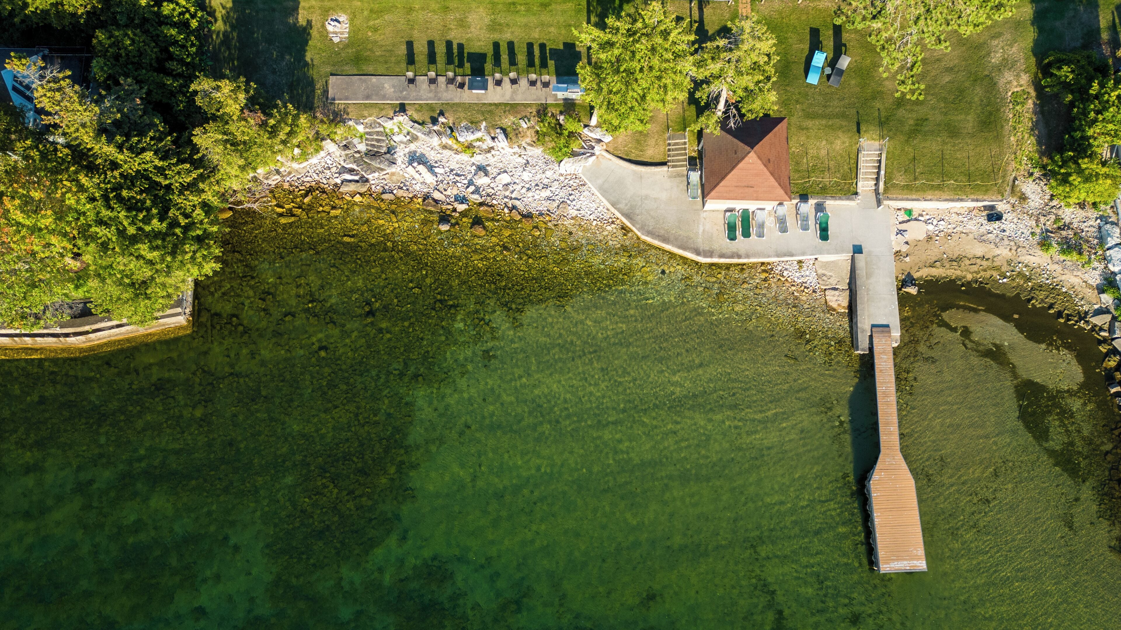Vista aérea de un parque junto al lago con un muelle de madera, un pequeño pabellón y una zona de césped con bancos.