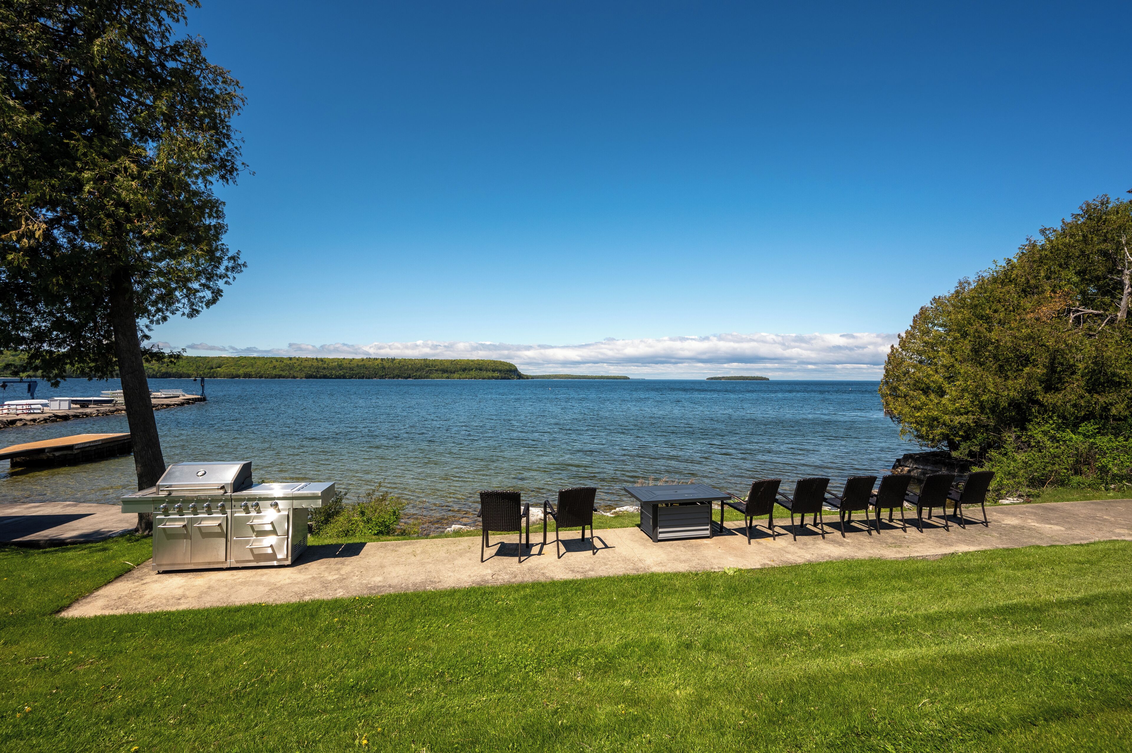 A lakeside patio with a grill, chairs, and a table.