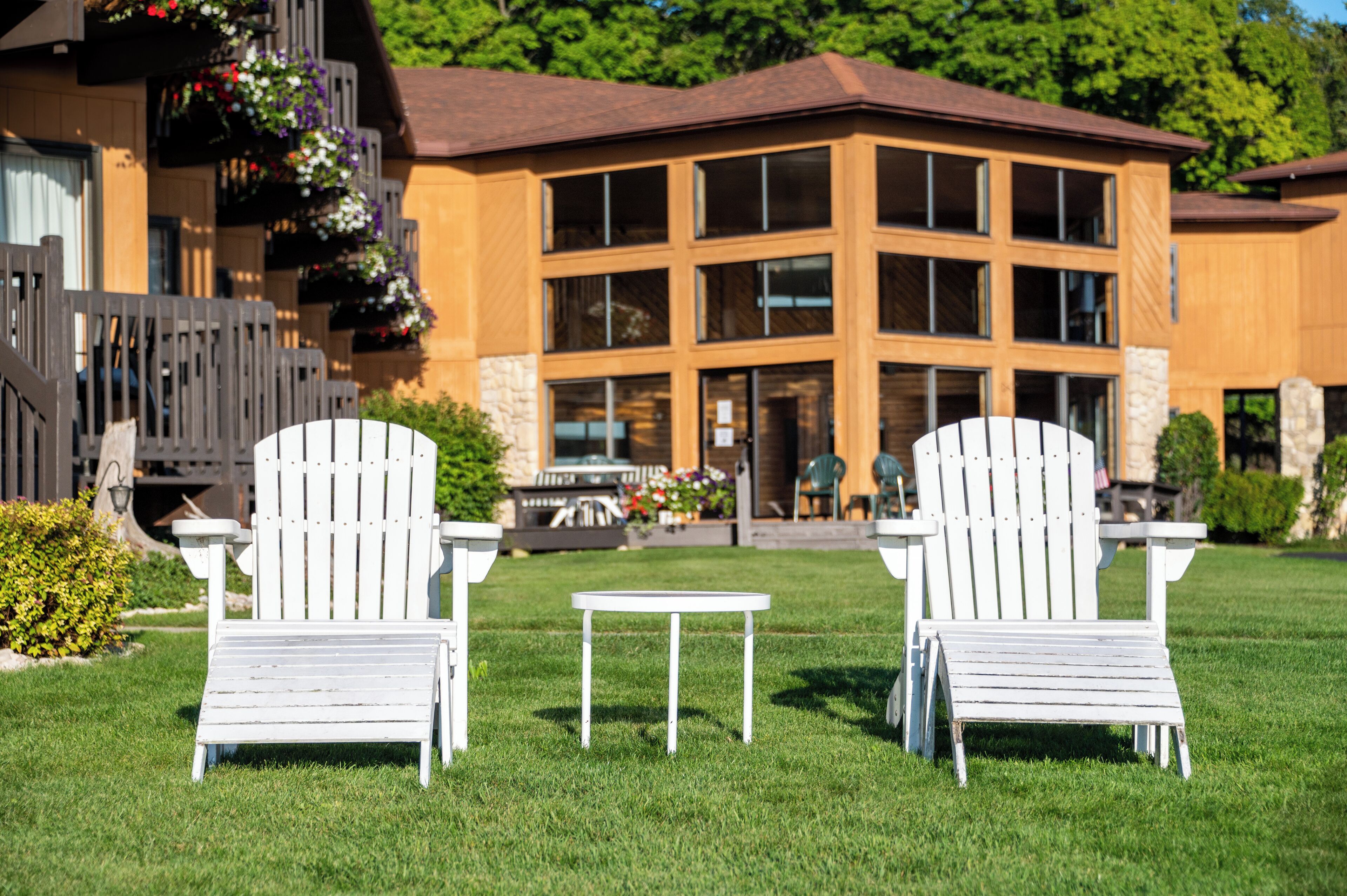 Two white Adirondack chairs with a small table between them, set on a well-maintained lawn in front of a building with large windows and a wooden facade.