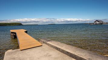Un muelle de madera se extiende sobre aguas tranquilas con un borde de concreto, una pequeña embarcación amarrada a la derecha e isla distante con un faro.