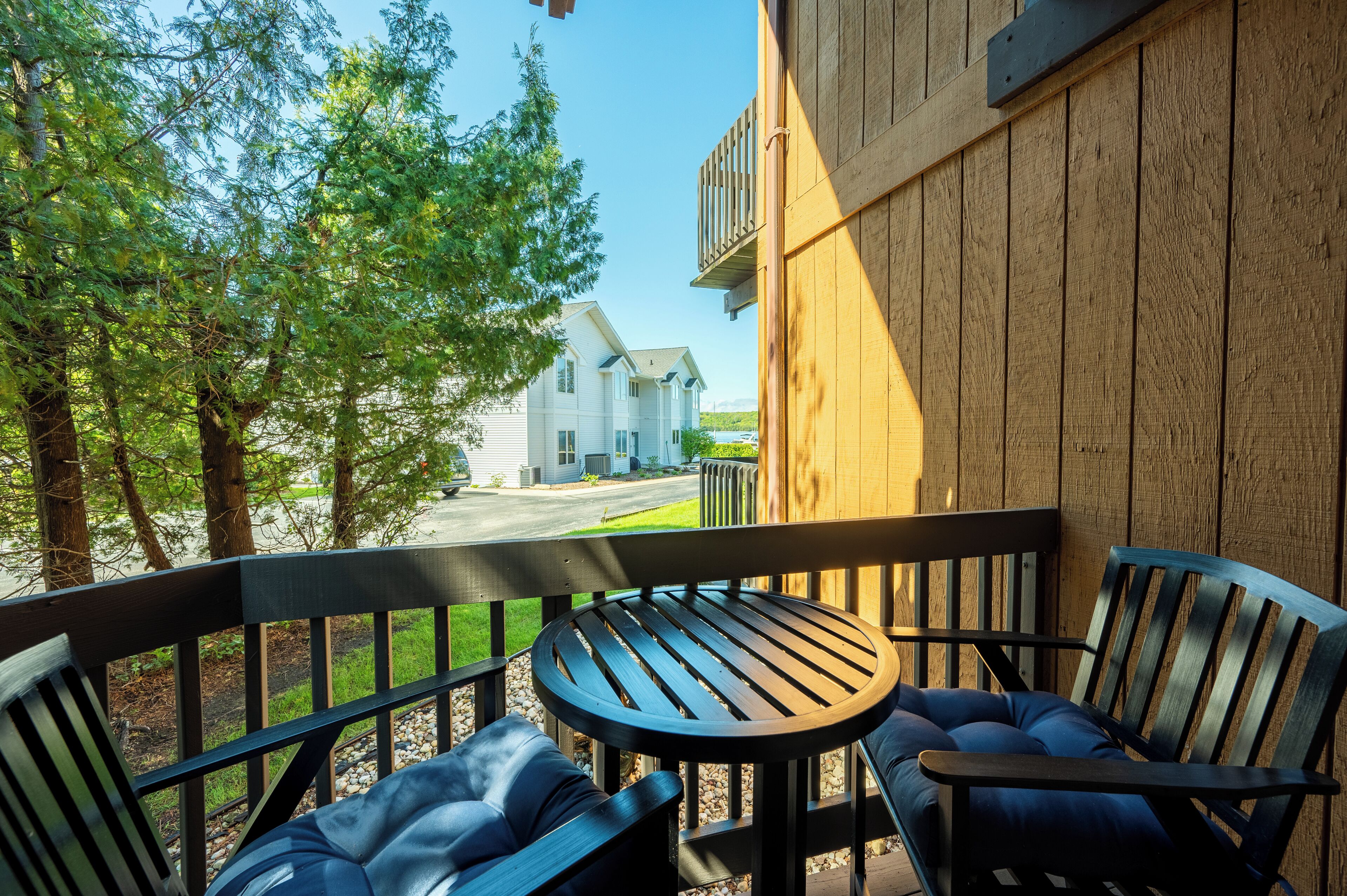 A wooden deck with a round table and two chairs, overlooking a road and a house.