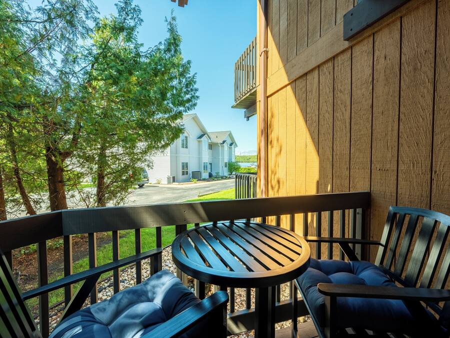A wooden deck with a round table and two chairs, overlooking a road and a house.